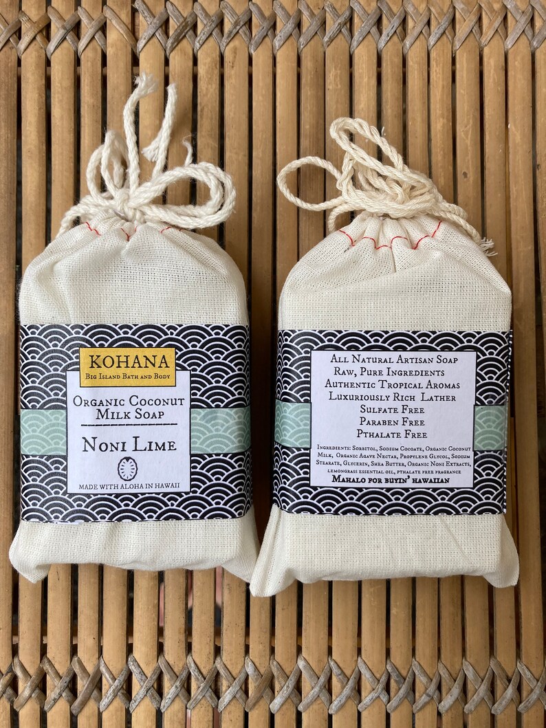 May include: Two bars of handmade soap wrapped in white muslin bags with red stitching. The soap on the left is labeled "KOHANA Big Island Bath and Body Organic Coconut Milk Soap Noni Lime Made with Aloha in Hawaii". The soap on the right is labeled "All Natural Artisan Soap Raw, Pure Ingredients Authentic Tropical Aromas Luxuriously Rich Lather Sulfate Free Paraben Free Phthalate Free Ingredients: Sorbitol, Sodium Cocoate, Organic Coconut Milk, Organic Aloe Vera, Propylene Glycol, Sodium Stearate, Glycerin, Shea Butter, Organic Noni Extract, Lemongrass Essential Oil, Phthalate Free Fragrance Mahalo for Buyin' Hawaiian"