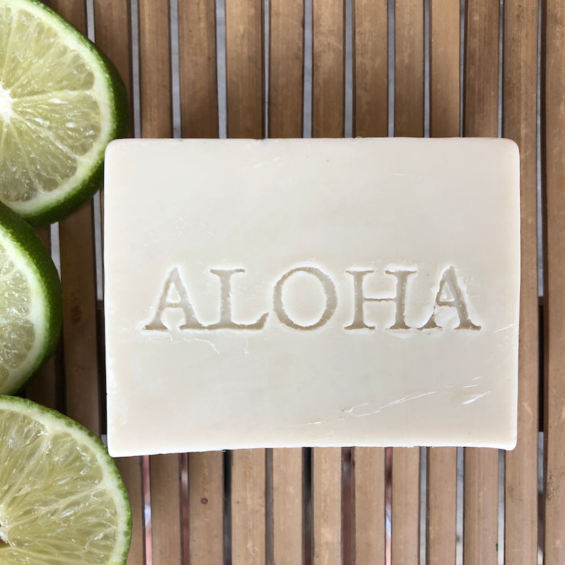 May include: A white bar of soap with the word "ALOHA" embossed on it. The soap is sitting on a bamboo mat with lime slices beside it.