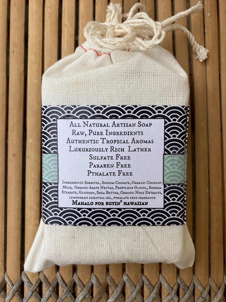May include: A white muslin bag with a drawstring closure containing a bar of natural artisan soap. The soap label features black and white waves and text that reads "All Natural Artisan Soap, Raw, Pure Ingredients, Authentic Tropical Aromas, Luxuriously Rich Lather, Sulfate Free, Paraben Free, Phthalate Free, Ingredients: Sorbitol, Sodium Cocoate, Organic Coconut Milk, Organic Agave Nectar, Propylene Glycol, Sodium Stearate, Glycerin, Shea Butter, Organic Noni Extracts, Lemongrass Essential Oil, Phthalate Free Fragrance, Mahalo for Buyin' Hawaiian."