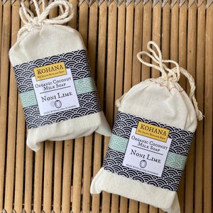 May include: Two white cotton drawstring bags containing bars of organic coconut milk soap. The soap is labeled "KOHANA Big Island Bath and Body" and "NONI LIME" with a green and black design. The bags are sitting on a woven bamboo mat.