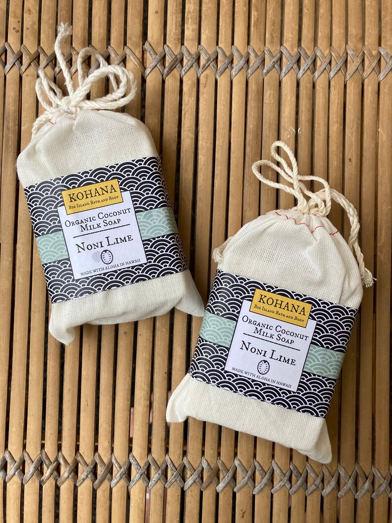 May include: Two white cotton drawstring bags containing bars of organic coconut milk soap. The soap is labeled "KOHANA Big Island Bath and Body" and "NONI LIME" with a green and black design. The bags are sitting on a woven bamboo mat.
