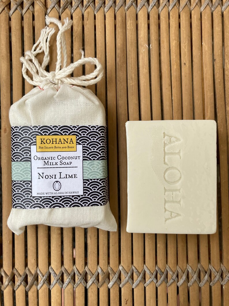May include: A white bar of soap with the word "ALOHA" embossed on it. The soap is packaged in a white muslin bag with a black and white label that reads "KOHANA Big Island Bath and Body Organic Coconut Milk Soap Noni Lime Made with Aloha in Hawaii".