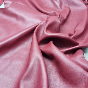 May include: Close-up of a sheet of burgundy leather with a smooth, shiny surface. The leather is folded and draped, showing the texture and color variations. The edges of the leather are visible, revealing the thickness and quality of the material.