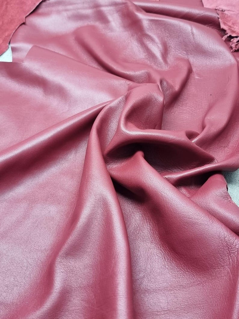 May include: Close-up of a sheet of burgundy leather with a smooth, shiny surface. The leather is folded and draped, showing the texture and color variations. The edges of the leather are visible, revealing the thickness and quality of the material.