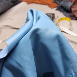 以下が含まれることがあります： 柔らかく、質感のある表面を持つ、明るい青色の革のクローズアップ。革は折りたたまれ、より明るい青色の裏側が現れています。背景には、さまざまな色の革片とメジャーが写っています。