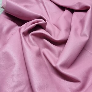 May include: A sheet of soft, pink leather with a textured surface. The leather is folded and draped, showing the natural grain and texture. The edges are slightly frayed, adding to the organic feel of the material. Ideal for crafting.