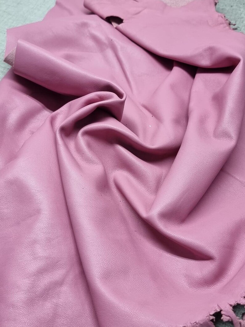 May include: A sheet of soft, pink leather with a textured surface. The leather is folded and draped, showing the natural grain and texture. The edges are slightly frayed, adding to the organic feel of the material. Ideal for crafting.