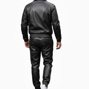 May include: A black leather jacket and matching pants set. The jacket has a collar and ribbed cuffs and waist. The pants have a ribbed ankle cuff. The person is wearing white sneakers.