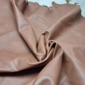 May include: A close-up shot of a folded piece of brown leather. The leather has a smooth, matte finish and a warm, earthy tone. The edges of the leather are uneven, suggesting it is a natural material. The folds create shadows and highlights, adding depth to the image.