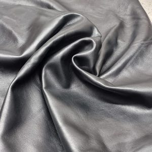 May include: Close-up of a sheet of black leather with a smooth, shiny surface. The leather is folded and wrinkled, creating shadows and highlights. The material appears soft and pliable, suitable for various crafting projects.