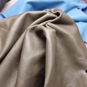 May include: Close-up of olive green leather fabric with soft folds and a smooth texture. The leather is the primary focus, with a light blue fabric visible in the background. The image highlights the material's quality and color.