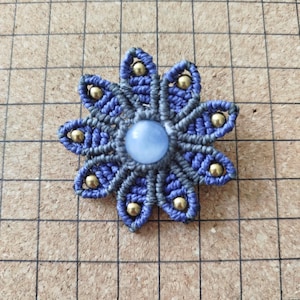 Peut inclure: Une fleur en macramé faite à la main avec huit pétales bleus et gris. Chaque pétale a une perle dorée à l'extrémité. Le centre présente une pierre ronde bleu clair. La fleur est posée sur un fond quadrillé marron.
