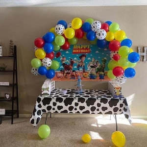 Peut inclure: D&eacute;coration de f&ecirc;te d'anniversaire avec une table recouverte d'une nappe &agrave; imprim&eacute; vache noir et blanc. Une arche de ballons rouges, jaunes, bleus, verts et &agrave; imprim&eacute; dalmatien. La toile de fond affiche le texte "HAPPY BIRTHDAY".