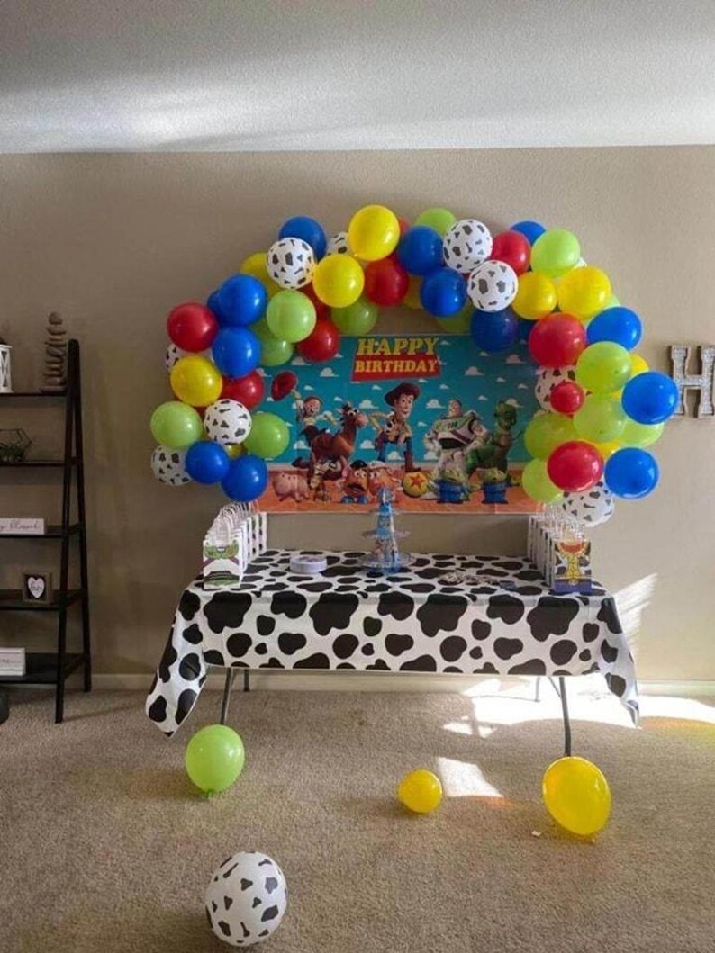 Peut inclure: D&eacute;coration de f&ecirc;te d'anniversaire avec une table recouverte d'une nappe &agrave; imprim&eacute; vache noir et blanc. Une arche de ballons rouges, jaunes, bleus, verts et &agrave; imprim&eacute; dalmatien. La toile de fond affiche le texte "HAPPY BIRTHDAY".