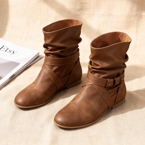 Pode incluir: Um par de botins castanhos com um design folgado. As botas apresentam biqueira redonda, uma tira com fivela e um salto baixo. A palavra "Etsy" é visível numa revista no fundo.