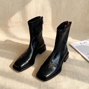 以下が含まれることがあります： 黒い革のアンクルブーツのペア。つま先は四角く、ローヒールです。ブーツの内側にはジッパーがあり、滑らかで光沢のある仕上がりです。ブーツはニュートラルな色の表面に置かれています。