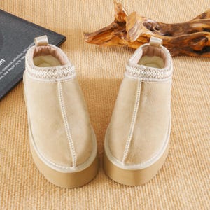 Könnte beinhalten: Ein Paar beige Wildleder-Schlupfschuhe mit einer dicken, hellbraunen Sohle. Die Schuhe haben einen cremefarbenen, gemusterten Besatz um den Knöchel und ein weiches, helles Innenfutter. Die Nähte sind in der Mitte des Schuhs sichtbar.