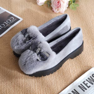 Pode incluir: Um par de mocassins cinzentos de camurça com um detalhe de pele sintética e um laço decorativo com strass. Os sapatos têm uma sola preta e um interior macio e felpudo. Estes mocassins são projetados para conforto e estilo.