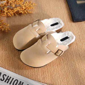 Pode incluir: Um par de tamancos de camurça bege com forro de pele sintética. Os tamancos têm fecho de fivela e sola de cortiça. Estes sapatos de interior oferecem conforto e calor.