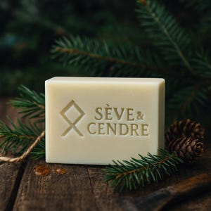 Peut inclure: Un pain de savon rectangulaire blanc cassé avec les mots "SÈVE & CENDRE" et un motif géométrique gravé. Le savon est posé sur une surface en bois avec des aiguilles de pin et des pommes de pin.
