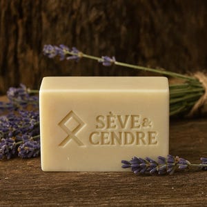 Peut inclure: Un savon rectangulaire beige clair avec les mots "Sève & Cendre" gravés, accompagnés d'un motif géométrique. Des brins de lavande sont disposés autour du savon. Le fond est une surface en bois sombre.