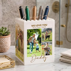 May include: A white pen holder with a collage of family photos and the words "WE LOVE YOU" and "Daddy" in gold script. The holder contains five pens in black, green, gold, silver, and navy blue.
