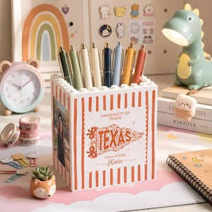 May include: A white and orange striped pen holder with the text "UNIVERSITY OF TEXAS TEXAS Class of 2026 Katie" holds several pens. A green dinosaur lamp, a pink and blue clock, and decorative items are also visible.