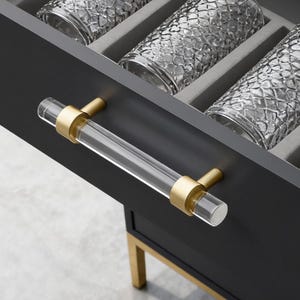 May include: A dark gray cabinet drawer is open, revealing four clear, textured drinking glasses nestled in a gray foam insert. The drawer features a clear acrylic handle with gold-colored accents. The cabinet has gold-colored legs.