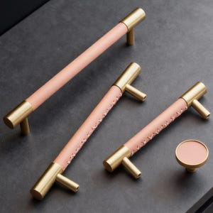 May include: A collection of cabinet handles and a knob. The handles are made of brass with a light pink leather wrap, stitched with a contrasting thread. The knob is round and matches the handle's design. The hardware is designed for cabinets.