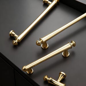 May include: A collection of gold-colored cabinet hardware, including long bar pulls and shorter handles. The hardware is arranged on a dark surface, showcasing the polished finish and decorative end details. The pulls vary in size, offering design options.