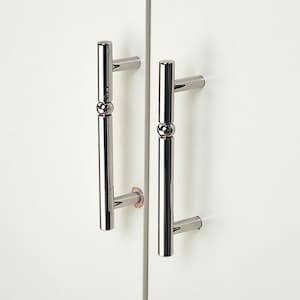 May include: Close-up of two polished chrome cabinet door handles. Each handle has a cylindrical body with a decorative sphere in the middle and a rectangular grip at each end. The handles are mounted on a white cabinet door.