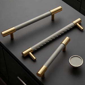 Peut inclure: Ensemble de quincaillerie d'armoire comprenant des poignées en laiton et en cuir gris, ainsi qu'un bouton assorti. Les poignées sont cylindriques avec des embouts et des montants en laiton. Le cuir gris est cousu au centre de certaines poignées.