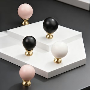 Könnte beinhalten: Eine Auswahl an dekorativen Schrankknöpfen in Schwarz, Weiß und Rosa, jeweils mit einem goldfarbenen Sockel. Die Knöpfe sind auf einer weißen geometrischen Plattform vor dunklem Hintergrund präsentiert.