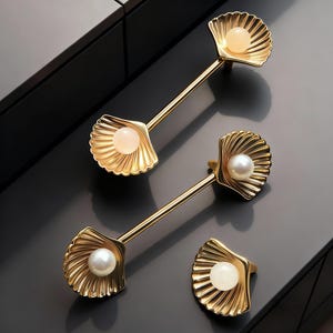 Peut inclure: Poignées de meuble dorées avec un design de coquillage. Chaque poignée présente un globe perlé au centre de la coquille. Les poignées sont montées sur une surface gris foncé, contrastant avec la finition dorée.