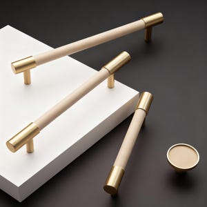 Puede incluir: Un juego de herrajes para armarios con tiradores de madera clara y detalles metálicos color latón. El juego incluye tiradores largos y un pomo redondo. Los artículos están dispuestos sobre una superficie blanca sobre un fondo negro.