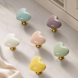 May include: A collection of six heart-shaped cabinet knobs in pastel colors including light blue, mauve, cream, peach, sage green, and yellow. Each knob features a smooth, glossy finish and is attached to a circular brass-toned metal base.