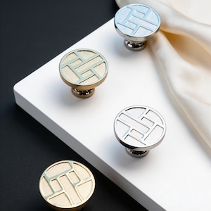 Op de afbeelding: Vier decoratieve kastknoppen met een geometrisch ontwerp. De knoppen hebben een cirkelvormig oppervlak met een verzonken patroon in lichtblauw, zilver en goud. De knoppen liggen op een wit oppervlak met een zwarte achtergrond.