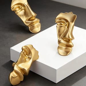 Può includere: Tre sculture dorate raffiguranti volti con un design moderno e astratto. Le sculture sono esposte su una piattaforma bianca e una superficie grigio scuro. I volti hanno una finitura metallica liscia e sono disposti in modo accattivante.