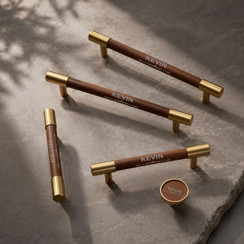 May include: Three long cylindrical cabinet pulls and one small round cabinet knob, each featuring a brown leather-wrapped centre with gold metal ends. The word KEVIN is printed in white on the leather surface of every piece in this hardware set.