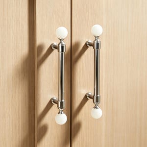 May include: Two cabinet door pulls with a polished chrome finish and white spherical accents. The pulls are vertically oriented and mounted on light-colored wooden cabinet doors. The design is modern and elegant.