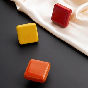 May include: Three square, glossy objects in yellow, red, and orange are arranged on a black surface with a white satin fabric. The objects have rounded edges and a reflective surface, creating a modern and minimalist aesthetic.