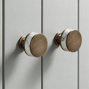 May include: Two cabinet knobs with a brushed brass finish and a white marble accent band. The knobs are round and mounted on a light gray cabinet door. The hardware has a classic, elegant design.