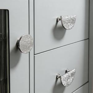 May include: A light grey cabinet with three drawers, each fitted with silver drawer pulls. One is a circular knob with a textured finish, and the other two are semi-circular with a textured surface.