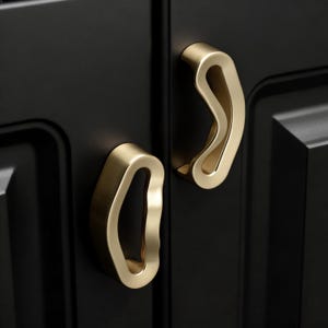 Op de afbeelding: Close-up van twee goudkleurige kastgrepen met een uniek, gebogen ontwerp. De grepen zijn gemonteerd op een zwarte kastdeur, wat een opvallend contrast creëert. De grepen hebben een gladde, gepolijste afwerking.