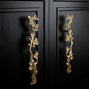 May include: Two gold-colored cabinet pulls in the shape of flowering branches. The pulls are mounted on a black cabinet door. The floral design adds a touch of elegance to the hardware.