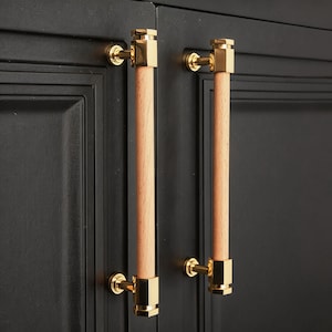 May include: Two cabinet pulls with a gold finish and light wood center. The pulls are mounted on a black cabinet door. The gold hardware has a square design at each end of the wood handle. The cabinet door has a recessed panel design.
