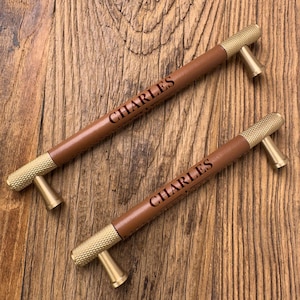 May include: Two drawer pulls featuring a brown leather-wrapped centre section and textured gold-tone metal ends. The leather portion of each handle is engraved with the name CHARLES in a dark, clear font, centred between two horizontal lines.
