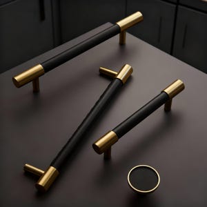 Peut inclure: Ensemble de quincaillerie d'armoire avec finitions noires et dorées. L'ensemble comprend plusieurs barres de tirage et un bouton rond. Les barres ont un centre noir avec des extrémités dorées. Le bouton est noir avec un bord doré.