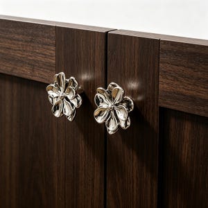 May include: Two silver flower-shaped cabinet knobs are attached to a dark brown wooden cabinet door. The knobs have a detailed petal design and a shiny, reflective surface. The wood grain is visible on the cabinet doors.