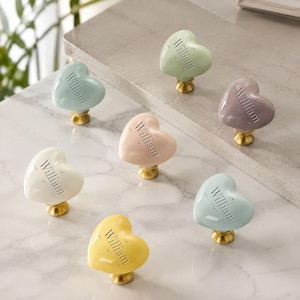 Puede incluir: Siete pomos para armarios en forma de corazón en varios colores pastel, incluyendo menta, rosa pálido, amarillo, blanco y lavanda. Cada pomo presenta el nombre William grabado en una fuente serif y una base de metal tono latón.